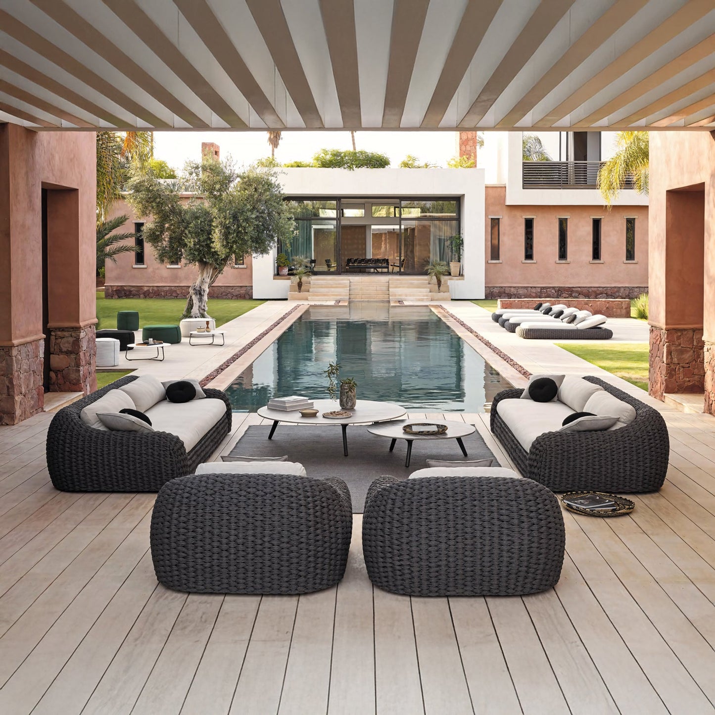 A spacious patio showcases the Palermo Terra Collection by Landmark Decor, featuring modern wicker furniture with all-weather frames and weather-resistant cushions. It faces a long rectangular pool, bordered by green lawns and palm trees, with a contemporary house and large glass windows overlooking the scene.