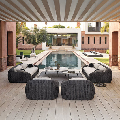 A spacious patio showcases the Palermo Terra Collection by Landmark Decor, featuring modern wicker furniture with all-weather frames and weather-resistant cushions. It faces a long rectangular pool, bordered by green lawns and palm trees, with a contemporary house and large glass windows overlooking the scene.