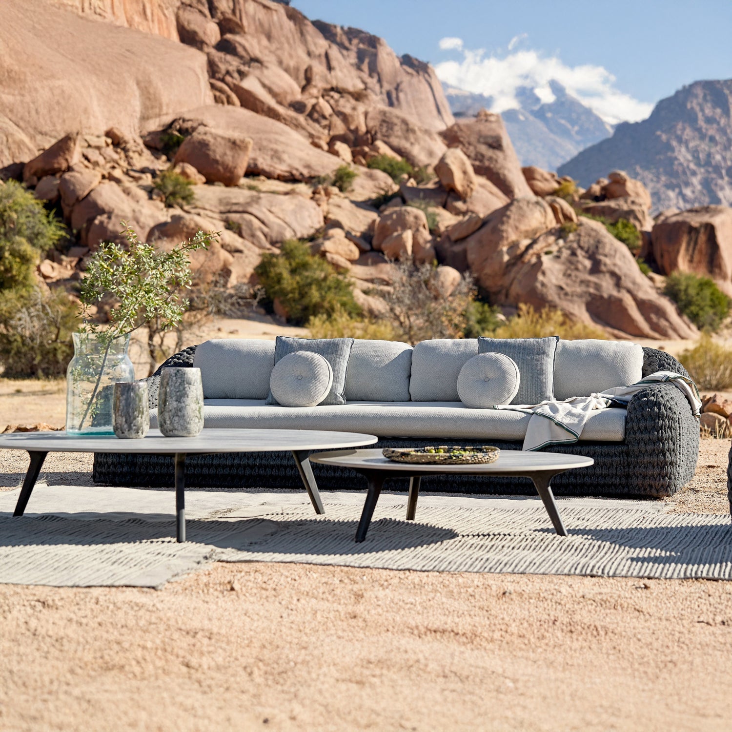 The Palermo Terra Collection by Landmark Decor features a woven grey sofa with weather-resistant cushions, circular coffee tables on a textured mat, and decorative vases and bowls. This outdoor seating area is elegantly set against a rocky desert landscape with mountains in the background.