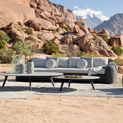 The Palermo Terra Collection by Landmark Decor features a woven grey sofa with weather-resistant cushions, circular coffee tables on a textured mat, and decorative vases and bowls. This outdoor seating area is elegantly set against a rocky desert landscape with mountains in the background.