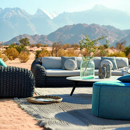The Palermo Terra Collection by Landmark Decor features a durable outdoor lounge setup, including a wicker sofa and ottoman with all-weather frames on a textured rug. A round table with foliage complements the desert landscape backdrop under a clear sky, enhanced by weather-resistant cushions.