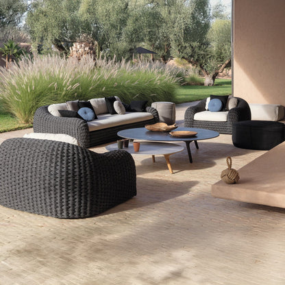 The Palermo Terra Collection by Landmark Decor features weather-resistant wicker outdoor sofas with cushions surrounding a round coffee table, set in a serene atmosphere shaded by trees and bordered by tall grass, all supported by all-weather frames.