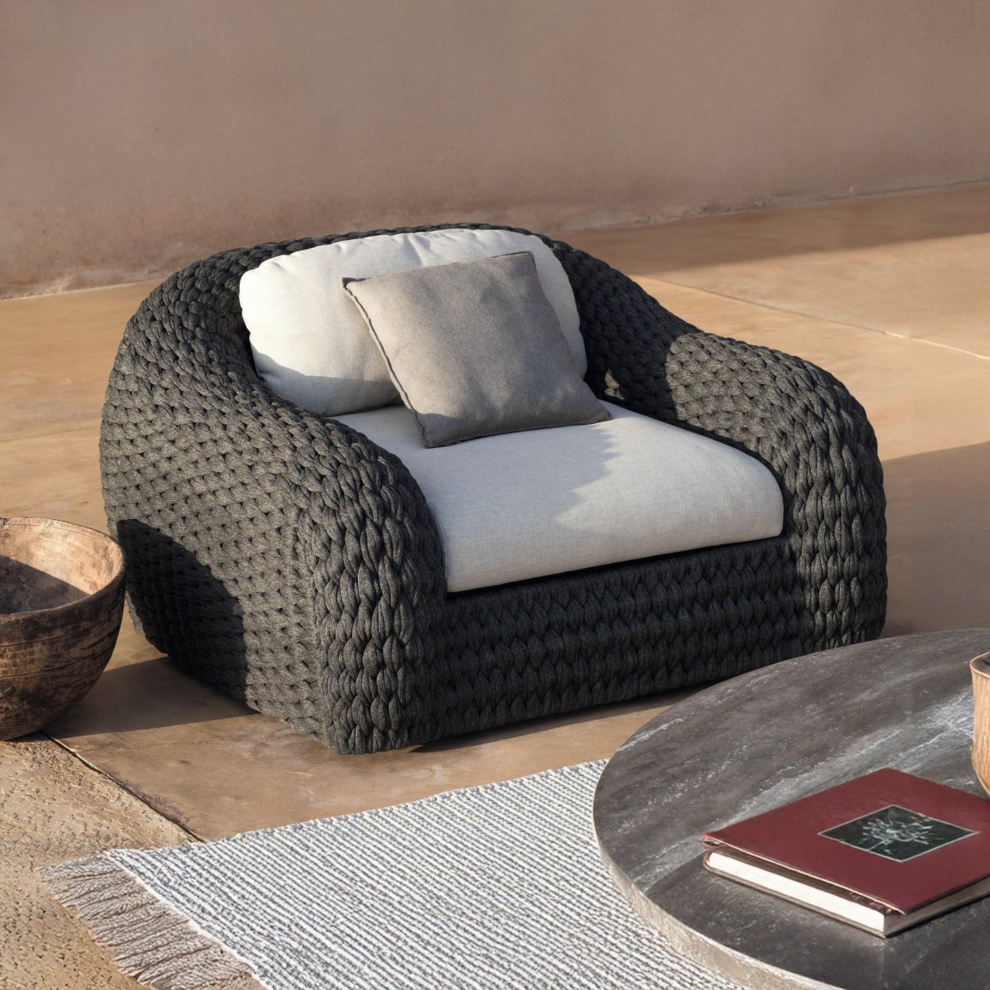 The Palermo Terra Collection by Landmark Decor features a woven dark gray armchair with weather-resistant cushions and a small pillow, complemented by a round stone table with books, a woven bowl, and a beige rug with white fringes. The all-weather frames make it perfect for outdoor use.