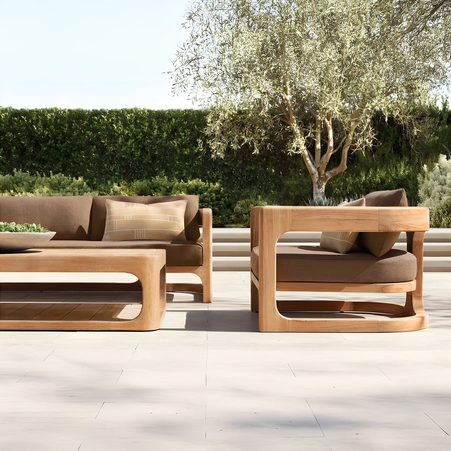 The Serene Arc Collection by Landmark Decor features a modern outdoor patio setting with wooden furniture and all-weather frames. It includes an outdoor sofa, chair, and coffee table with weather-resistant brown cushions, set against an olive tree and lush green hedge backdrop under a clear sky.