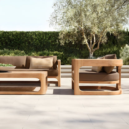The Serene Arc Collection by Landmark Decor features a modern outdoor patio setting with wooden furniture and all-weather frames. It includes an outdoor sofa, chair, and coffee table with weather-resistant brown cushions, set against an olive tree and lush green hedge backdrop under a clear sky.