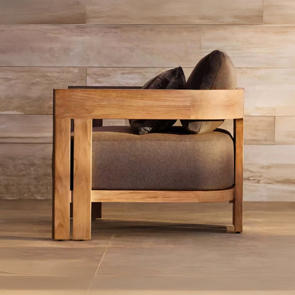 The Golden Horizon Collection by Landmark Decor showcases a wooden armchair with a boxy frame against a textured stone wall. It has weather-resistant gray fabric cushions and matching pillows, sitting elegantly on a light floor, perfect for indoor or outdoor settings.