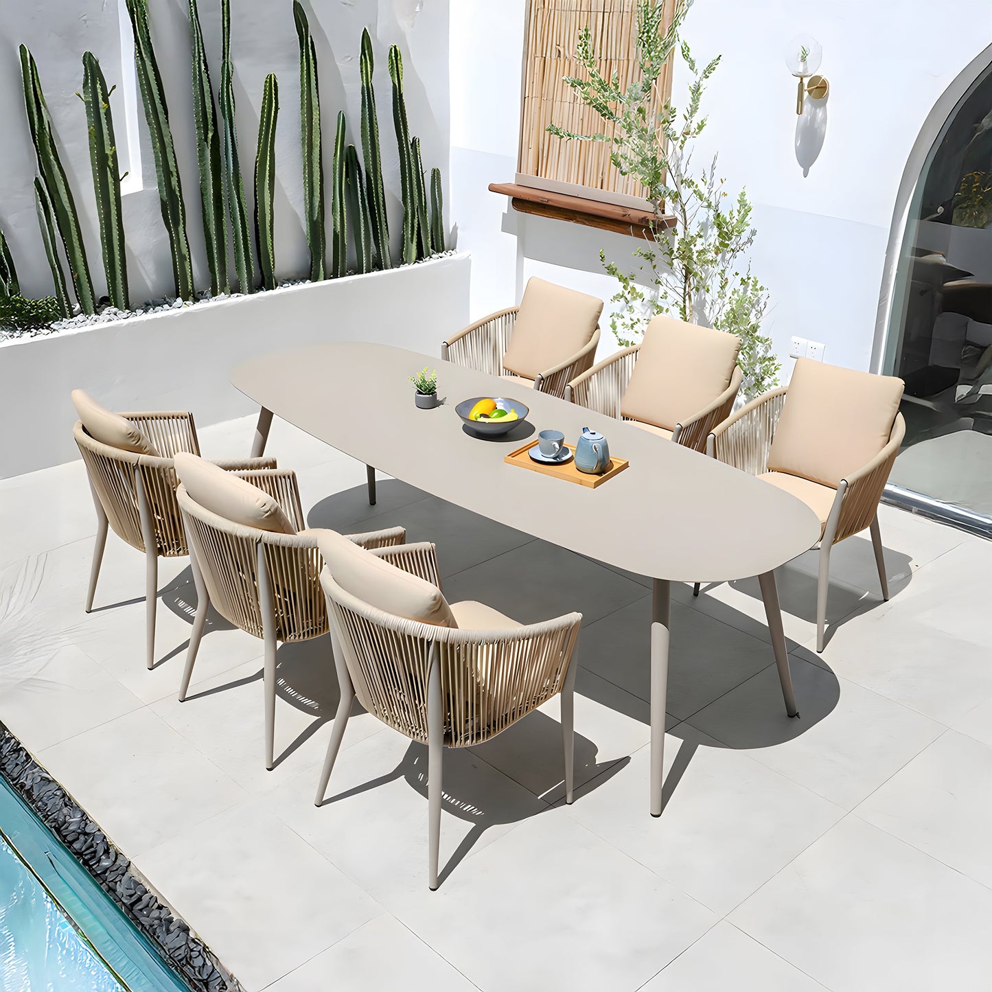The outdoor patio, ideal for alfresco dining, showcases The St. Tropez Collection by Landmark Decor, including a long oval table set for four with beige woven weather-resistant chairs. Adorned with a teapot and fruit bowls, its surrounded by cacti and a small tree on tiled flooring next to a serene water feature.