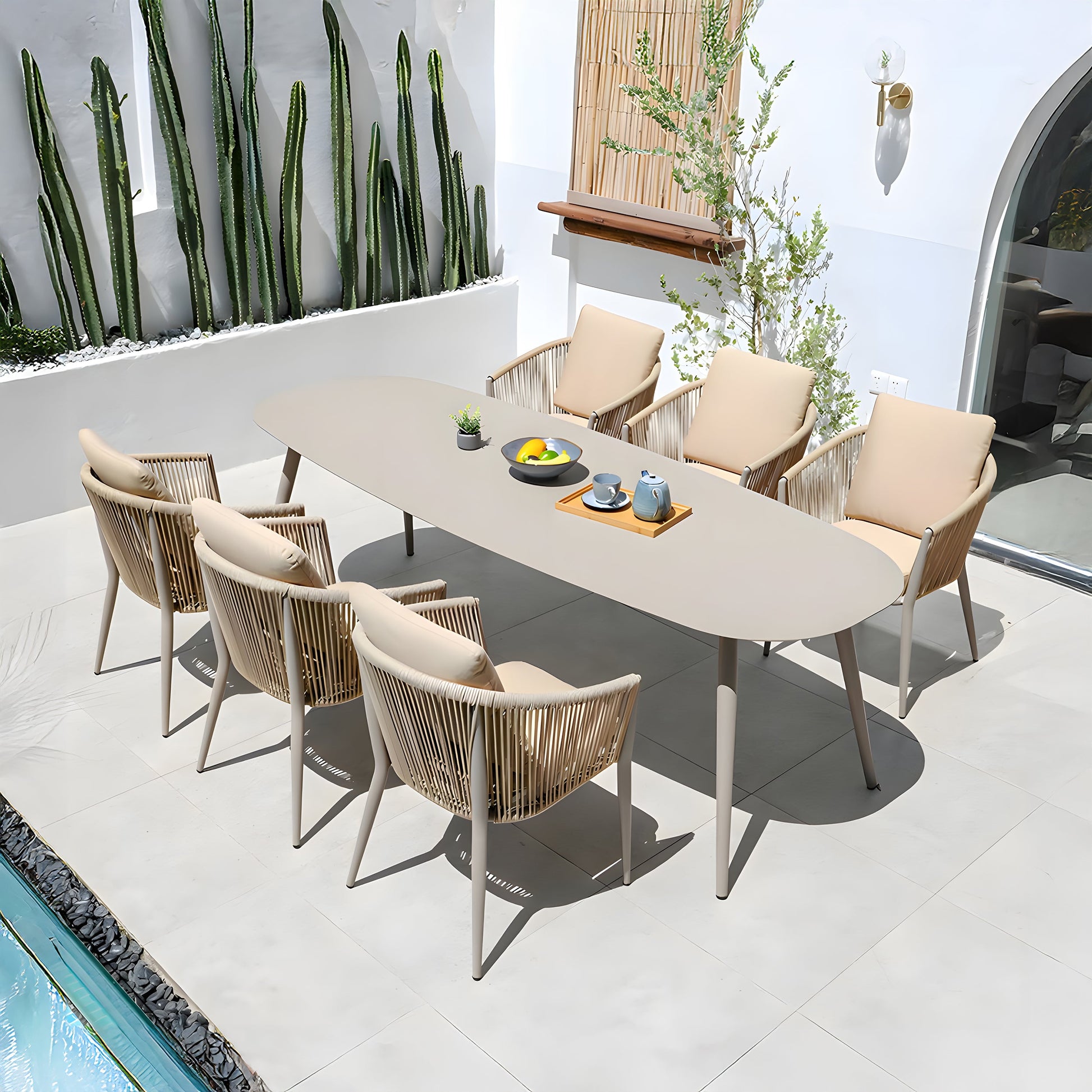 The outdoor patio, ideal for alfresco dining, showcases The St. Tropez Collection by Landmark Decor, including a long oval table set for four with beige woven weather-resistant chairs. Adorned with a teapot and fruit bowls, its surrounded by cacti and a small tree on tiled flooring next to a serene water feature.