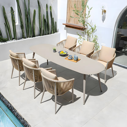 The outdoor patio, ideal for alfresco dining, showcases The St. Tropez Collection by Landmark Decor, including a long oval table set for four with beige woven weather-resistant chairs. Adorned with a teapot and fruit bowls, its surrounded by cacti and a small tree on tiled flooring next to a serene water feature.
