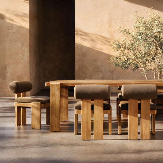 The Grove Retreat Collection by Landmark Decor offers a minimalist dining room setup with a large wooden table and five chairs made from weather-resistant materials, boasting sleek geometric lines. Sunlight casts gentle shadows, enhancing the textured backdrop adorned with a small tree in the corner.