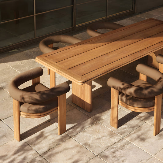 The Amber Ridge Collection by Landmark Decor, featuring a wooden dining table with four cushioned chairs, graces a sunlit stone patio. Shadows of trees dance over this weather-resistant set, creating an inviting alfresco dining experience.