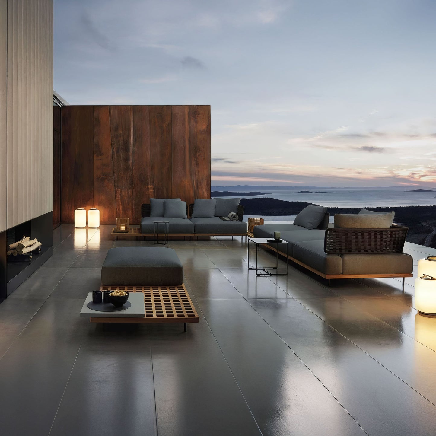 The Oceanview Oasis Collection by Landmark Decor offers sleek gray seating with wooden tables against a wooden accent wall. Weather-resistant cushions and lanterns complete the contemporary look, creating a serene setting with scenic sunset views over hills and water.