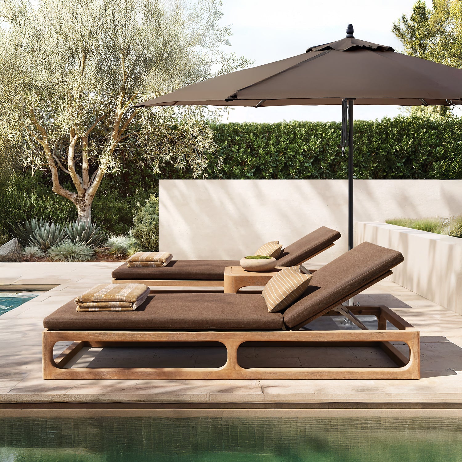 Beside the pool, two brown chaise lounges with pillows from Landmark Decors Bali Luxe Retreat Set sit under a large beige umbrella. A small wooden table crafted from premium materials holds a plant, while lush greenery and a stone patio complete the outdoor living space.