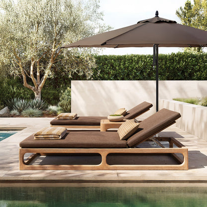 Beside the pool, two brown chaise lounges with pillows from Landmark Decors Bali Luxe Retreat Set sit under a large beige umbrella. A small wooden table crafted from premium materials holds a plant, while lush greenery and a stone patio complete the outdoor living space.