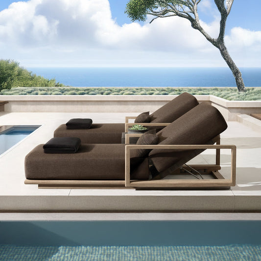 The patio features the Elysian Summit Lounger Set by Landmark Decor, complete with black pillows, beside a shimmering pool. A potted plant adds to the luxurious retreat vibe amidst an ocean view and partly cloudy skies, with a solitary tree framing the scene on the right.