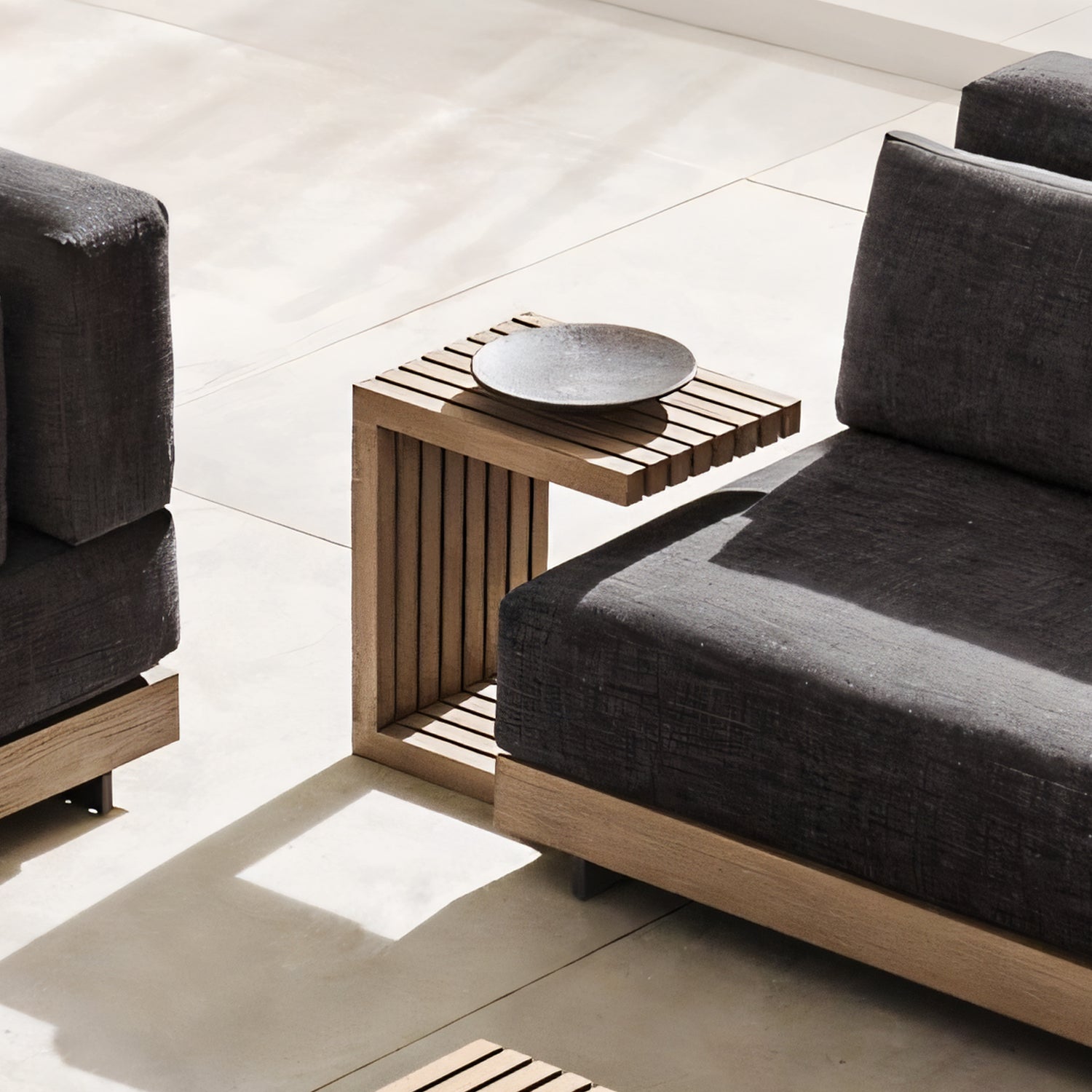A modern living room transforms into a luxurious retreat with the Golden Coast Lounger Set by Landmark Decor, featuring a dark gray sectional. A wooden side table with vertical slats holds a round black plate. Sunlight dances on the light-colored floor, enhancing the serene ambiance.