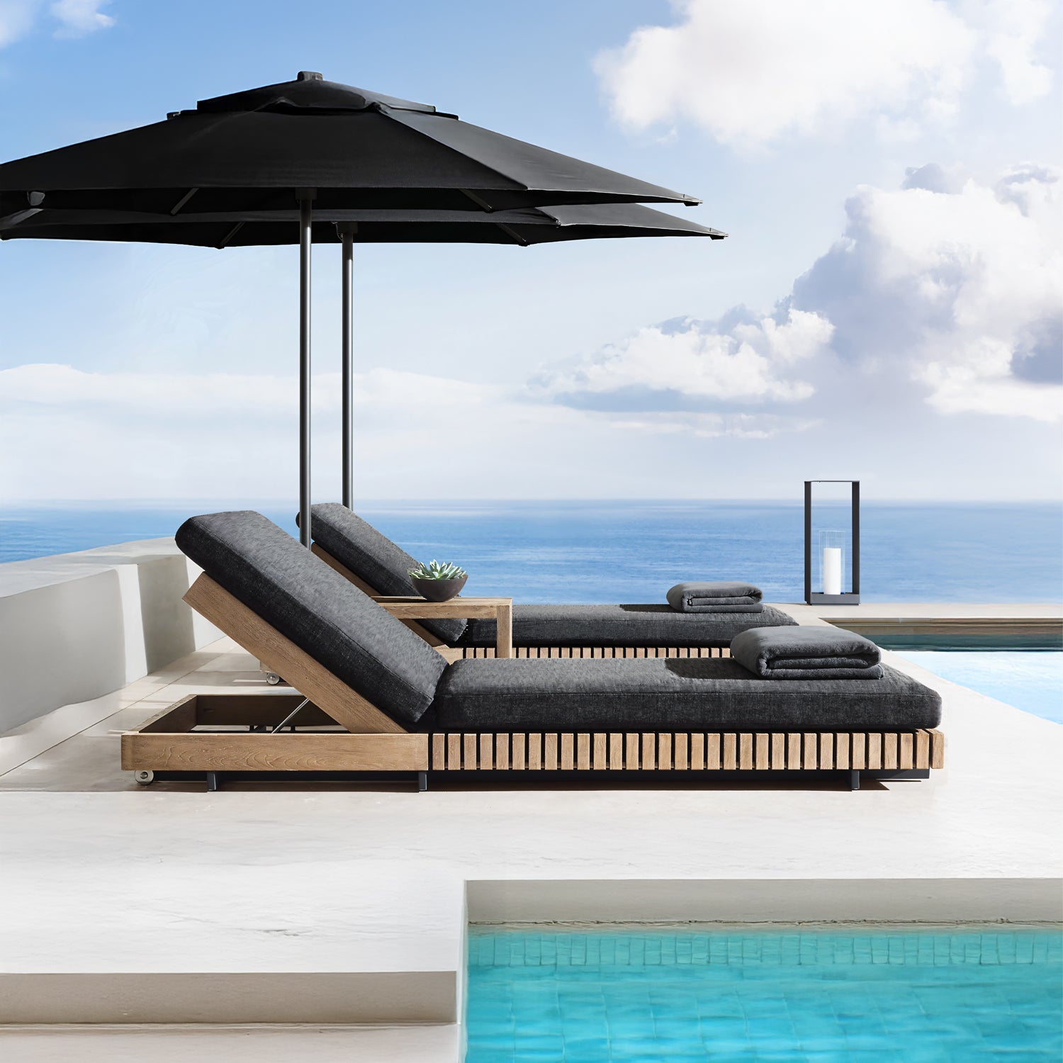 Experience luxury at the modern outdoor poolside, featuring Landmark Decors Golden Coast Lounger Set with two black-cushioned chaise lounges beneath a large umbrella. Overlooking the ocean with a clear blue sky, this serene retreat invites you to indulge in elevated outdoor living.