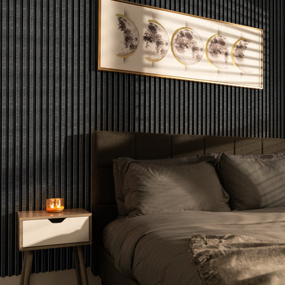 A modern bedroom features a dark accent wall with Landmark Decor's Obsidian Gridstone WPC Fluted Slat Wall Panel, a bed with gray bedding, a wooden nightstand holding a lit candle, and moon phase artwork above the headboard.