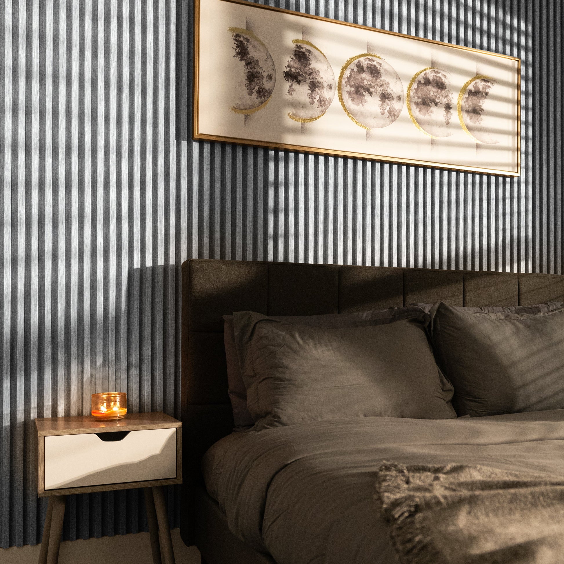 A neatly made bed with gray bedding sits by a wooden nightstand holding a lit candle; above, moon phase art and Landmark Decor’s Cloud Veil WPC Fluted Slat Wall Panel create an inviting atmosphere as striped shadows fall across the room.