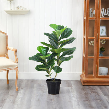 A medium Verdant Symphony plant by Landmark Decor graces the light wood floor, beside a cream chair and wooden cabinet with glass doors. A white paneled wall and decorative shelf backdrop enhance this inviting nook for the charming houseplant in its black pot.