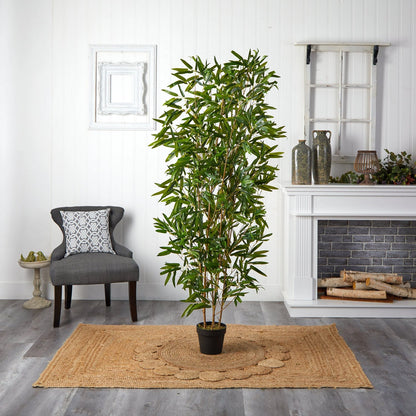 A cozy room features Landmark Decors Zen Whisper tall leafy plant on a woven rug. A gray armchair with a patterned cushion sits to the left, and the white mantelpiece to the right is adorned with decorative vases, while small logs nestle in the fireplace.