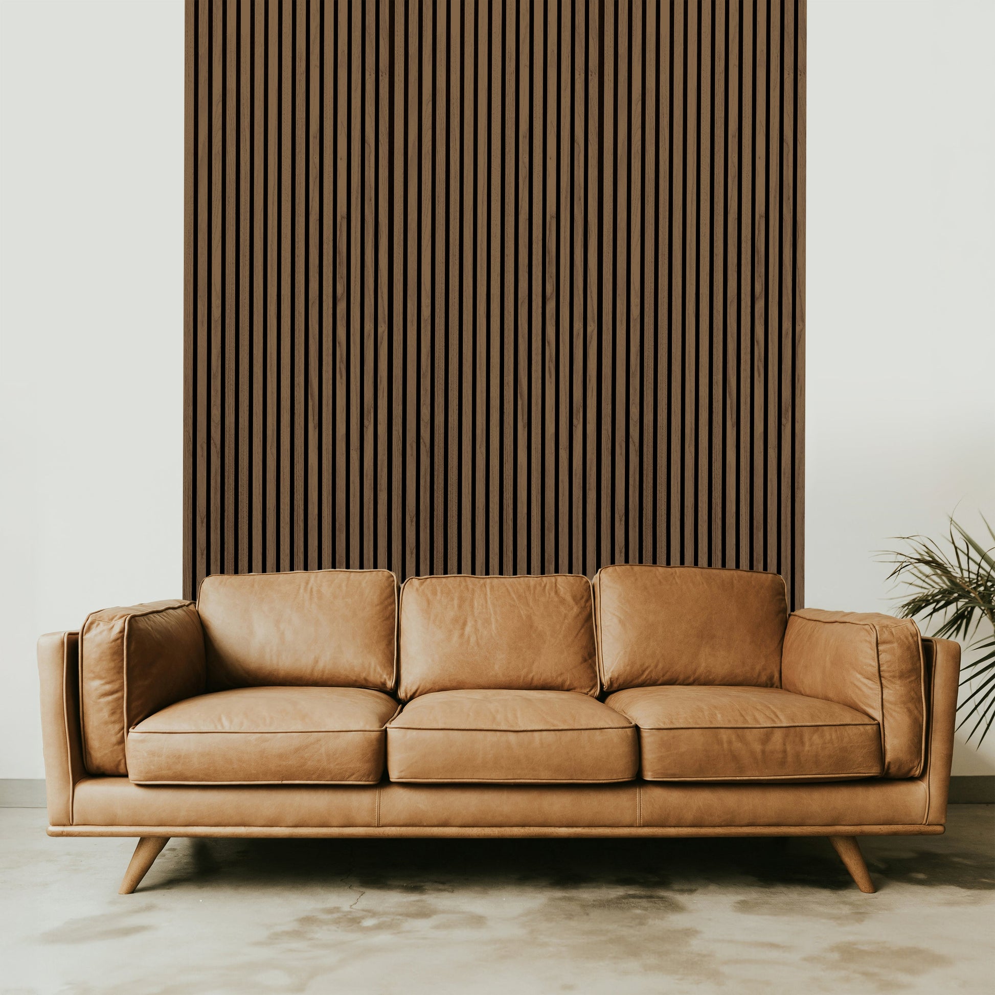 Tan leather sofa with three cushions in front of Landmark Decor’s Walnut Acoustic Slat Wall Panel, creating an elegant interior; a plant is partially visible on the right.